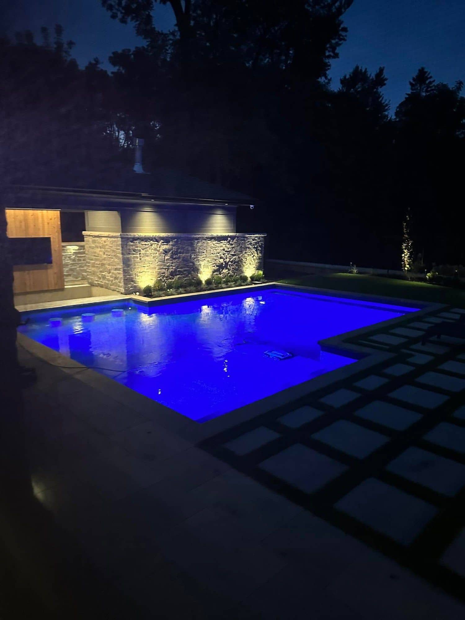 Inground pool lit blue at night beside a stone water wall