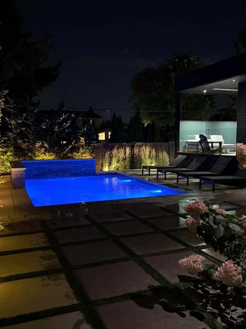 Custom inground pool lit blue at night beside a modern cabana and stone patio